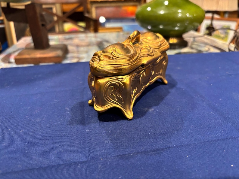 May include: A gold-colored jewelry box with a detailed, ornate design. The box has a curved lid with a sculpted figure on top. The box sits on four curved feet. The background is a blue surface.