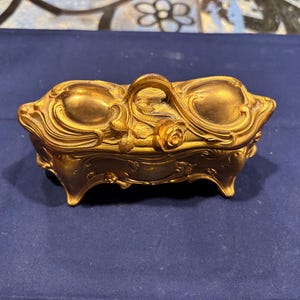 May include: A gold-colored, Art Nouveau style trinket box with a handle and floral details. The rectangular box has curved sides and sits on four small feet. The lid features a rose and other decorative elements.