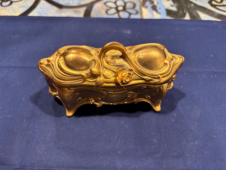 May include: A gold-colored, Art Nouveau style trinket box with a handle and floral details. The rectangular box has curved sides and sits on four small feet. The lid features a rose and other decorative elements.