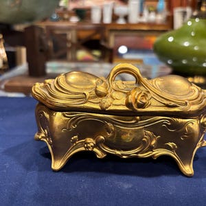 May include: Ornate gold-colored jewelry box with a curved lid and decorative floral accents. The box features a handle and rests on four curved feet. The design includes swirling patterns and a rose on the lid.