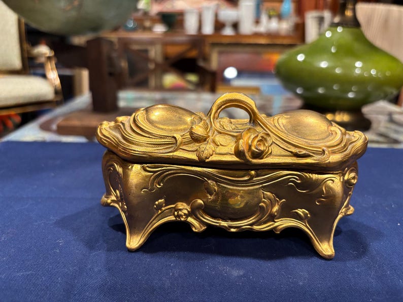 May include: Ornate gold-colored jewelry box with a curved lid and decorative floral accents. The box features a handle and rests on four curved feet. The design includes swirling patterns and a rose on the lid.