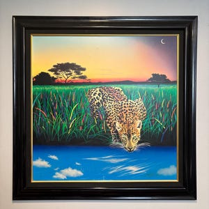 Peut inclure: Peinture encadrée d'un léopard buvant dans un plan d'eau bleu, sur fond de coucher de soleil. Le léopard a un pelage tacheté et est entouré de hautes herbes vertes. L'œuvre est dans un cadre noir.