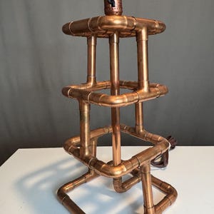 May include: A lamp base constructed from interconnected copper pipes and fittings. The structure features a multi-tiered, square-shaped frame with a central vertical pipe and a socket at the top. A brown electrical cord extends from the base.