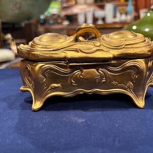 May include: A gold-colored, Art Nouveau style trinket box with a curved lid and ornate detailing. The box has a handle on top and sits on four curved feet. The box is made of metal and has a shiny finish. The box is on a blue surface.