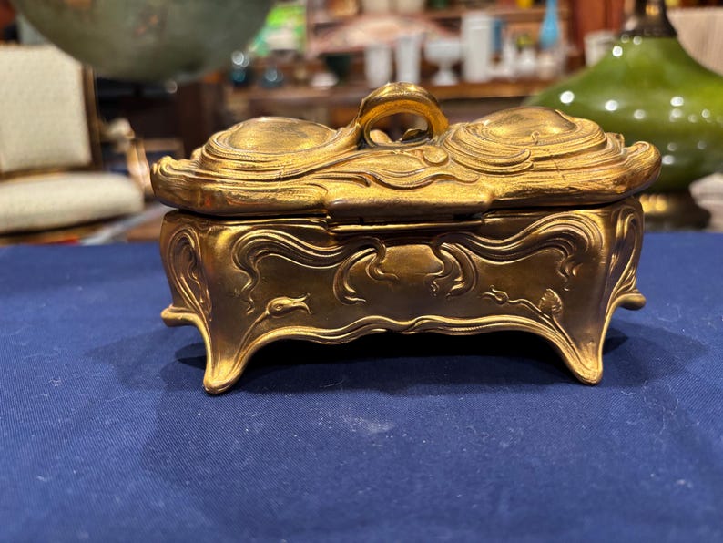May include: A gold-colored, Art Nouveau style trinket box with a curved lid and ornate detailing. The box has a handle on top and sits on four curved feet. The box is made of metal and has a shiny finish. The box is on a blue surface.