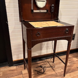 Puede incluir: Una mesa de tocador de caoba vintage con una tapa con bisagras que revela un espejo y una lámpara. La mesa tiene un cajón con un tirador de anillo y se apoya en patas delgadas. El cable eléctrico es visible.