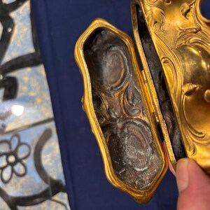 May include: An open, antique gold-colored jewelry box with ornate detailing. The interior is dark with a textured surface. The box is held open, revealing the inside. The background is a blue surface.