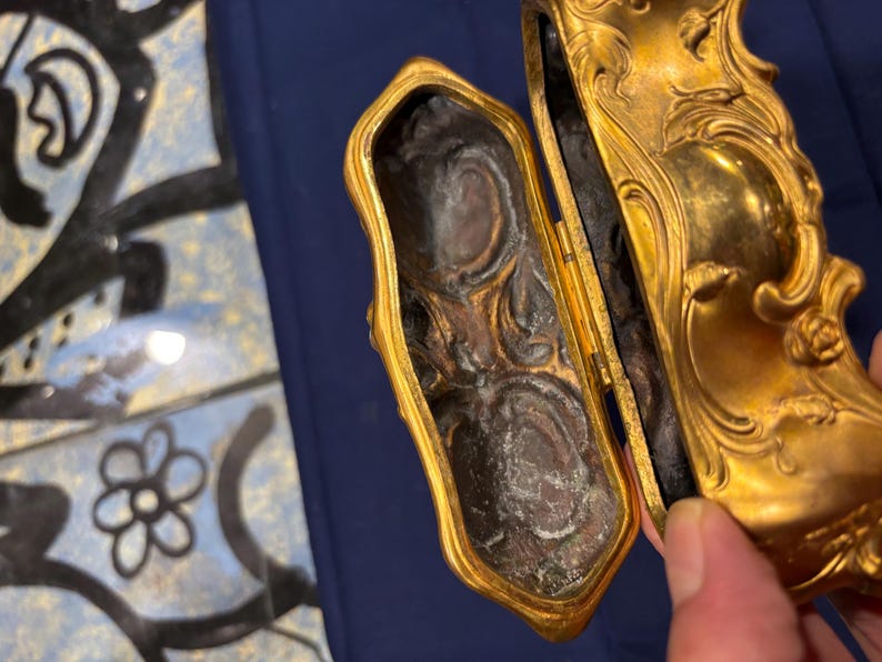 May include: An open, antique gold-colored jewelry box with ornate detailing. The interior is dark with a textured surface. The box is held open, revealing the inside. The background is a blue surface.
