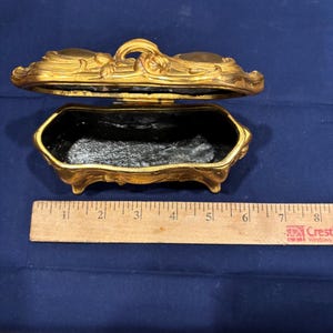 May include: An open, antique gold-colored jewelry box with a black interior. The box is rectangular with ornate floral detailing and rests on small feet. A wooden ruler is next to the box.