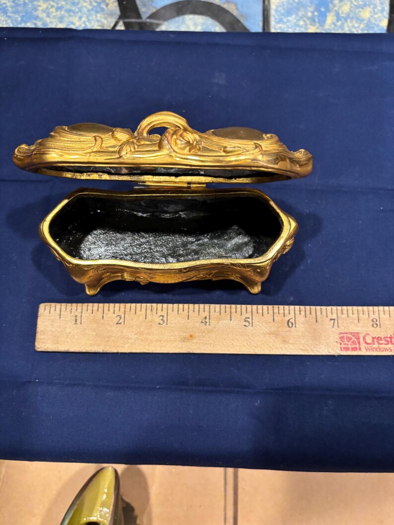 May include: An open, antique gold-colored jewelry box with a black interior. The box is rectangular with ornate floral detailing and rests on small feet. A wooden ruler is next to the box.