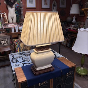 May include: A cream-colored table lamp with a pleated lampshade. The lamp has a decorative base and a brown wooden accent. The lamp is sitting on top of a speaker.