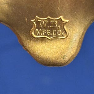 May include: Close-up of a gold-colored metal object with the text "W.B. MFG.CO" inside a decorative shield. The object has a smooth, curved shape and is set against a blue background.