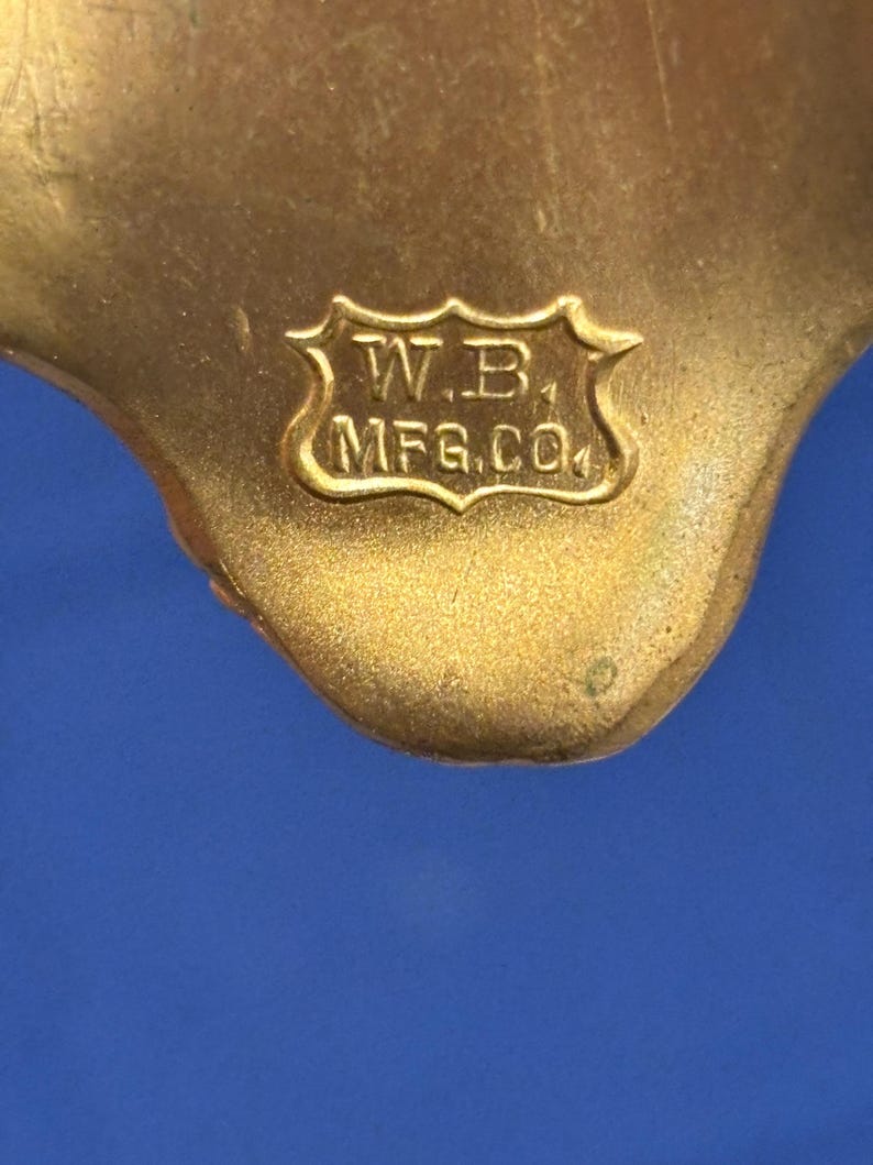 May include: Close-up of a gold-colored metal object with the text "W.B. MFG.CO" inside a decorative shield. The object has a smooth, curved shape and is set against a blue background.