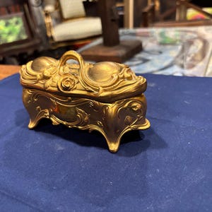May include: Ornate gold-colored jewelry box with a handle and decorative floral and scrollwork details. The box has four curved feet and sits on a blue surface. The lid features a rose design.