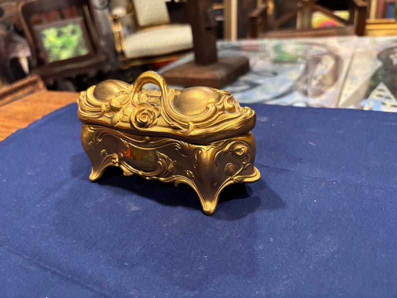 May include: Ornate gold-colored jewelry box with a handle and decorative floral and scrollwork details. The box has four curved feet and sits on a blue surface. The lid features a rose design.