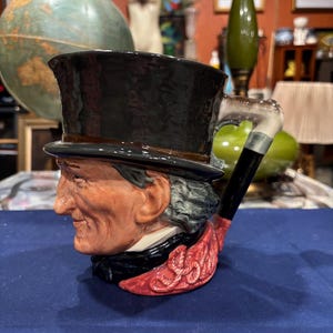 May include: A novelty ceramic mug depicting a man's head wearing a black top hat. The handle is a cane with a red and black design. The mug features a detailed face, black coat, and a red scarf.