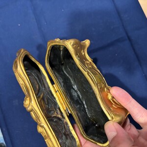 May include: An open, antique gold-colored box with ornate floral detailing. The interior is dark, and the box is held open, revealing the inside. The box has a hinged lid and a rectangular shape. The box is likely made of metal.