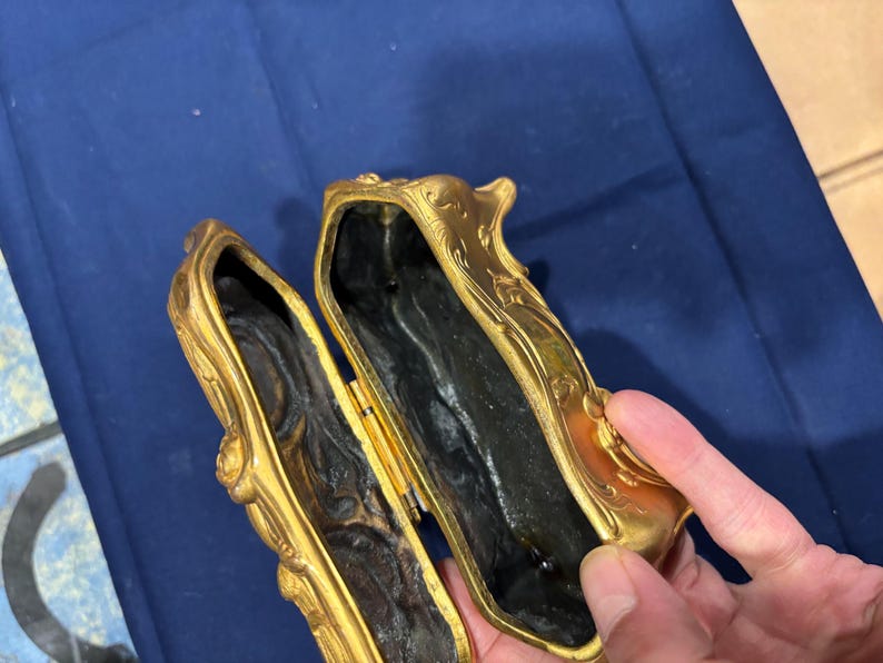 May include: An open, antique gold-colored box with ornate floral detailing. The interior is dark, and the box is held open, revealing the inside. The box has a hinged lid and a rectangular shape. The box is likely made of metal.
