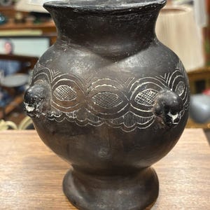 Op de afbeelding: Een donkergrijze, handgemaakte vaas met een bolvormig lichaam en een uitlopende rand. De vaas heeft ingewikkelde, wit geëtste patronen rond het middengedeelte en kleine, gestileerde handvatten. De vaas is waarschijnlijk gemaakt van keramiek of een vergelijkbaar materiaal.