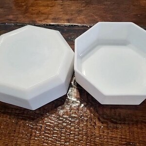 May include: Two white, octagonal ceramic dishes. One dish is shown upside down, displaying its smooth, flat base and angled sides, while the other is shown right side up, revealing its open, hollow interior and clean, white surface.