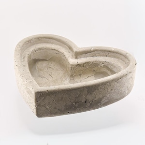 May include: A heart-shaped concrete planter in shades of gray. The planter has a layered design, with a deeper interior and a textured surface. The object is set against a white background, highlighting its form and texture.