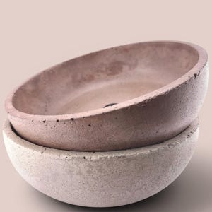 May include: Two stacked circular bowls with a rough, porous texture. The top bowl is a muted dusty rose color, while the bottom bowl is a lighter, off-white shade. Both bowls feature thick, rounded rims and a deep, concave interior basin.