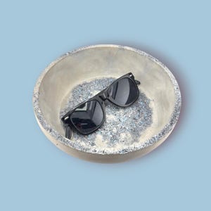 Könnte beinhalten: Eine runde, graue Betonschale mit strukturiertem Innenraum, gefüllt mit blauen und grauen Granulaten. Schwarze Sonnenbrille liegt in der Schale. Der Hintergrund der Schale ist hellblau.