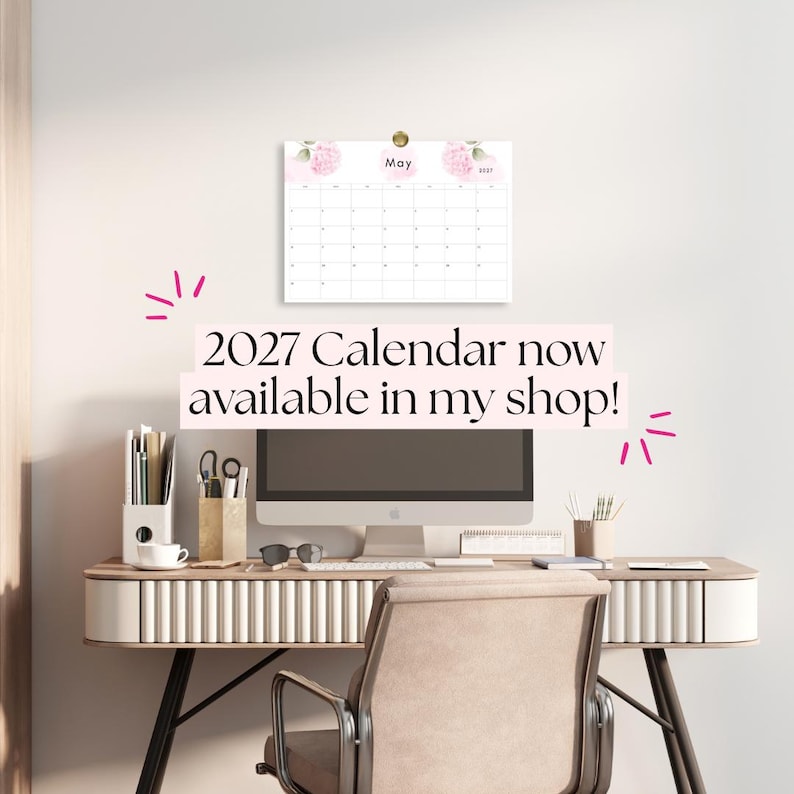 K&ouml;nnte beinhalten: Ein Wandkalender f&uuml;r 2027 mit einem Blumendesign und rosa Hortensien-Illustrationen am oberen Rand. Der Kalender zeigt den Monat Mai und ist &uuml;ber einem Computermonitor an der Wand befestigt, auf einem Schreibtisch mit B&uuml;robedarf.
