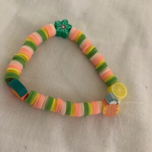 以下が含まれることがあります： ピンク、イエロー、グリーンの色の小さな丸いビーズで作られたカラフルなブレスレット。ブレスレットには、緑の花のチャーム、黄色のレモンのチャーム、パイナップルのチャームが付いています。