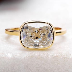 Op de afbeelding: Een gouden ring met een heldere, gefacetteerde edelsteen in kussenslijpvorm, gezet in een gladde zetting. De ringband is van dun, gepolijst goud en houdt de rechthoekige steen met afgeronde hoeken stevig op zijn plaats, met een glanzend oppervlak.