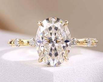Könnte beinhalten: Ein Goldring mit einem großen oval geschliffenen Mittelstein, der von vier Krappen gehalten wird. Die Ringschiene ist auf jeder Seite mit drei kleinen runden Steinen besetzt, die in einer Reihe angeordnet sind. Das Metall hat ein poliertes Finish.