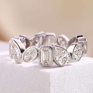 Può includere: Un anello color argento caratterizzato da una serie di pietre chiare alternate con taglio a pera, ovale e smeraldo, incastonate a lunetta. La fascia è contrassegnata con 925, indicando una composizione in argento sterling, con gemme sfaccettate.