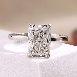 May include: A silver-toned ring featuring a large, rectangular radiant-cut center stone with multiple facets. The stone is held in place by four prongs on a smooth, polished metal band. The text Almira Jewels is visible on the band and corner.