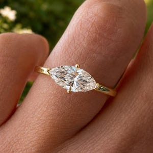 May include: A gold ring featuring a single marquise-cut clear gemstone set horizontally in a four-prong setting. The band is thin and polished, showcasing a simple design that highlights the elongated shape of the central stone.