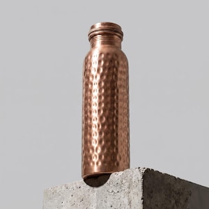 Peut inclure: Une bouteille d'eau cylindrique en cuivre présentant une texture martelée distinctive sur toute sa surface. La bouteille possède un goulot étroit et un bouchon à vis assorti avec un bord strié, affichant une finition brun-rouge métallique.