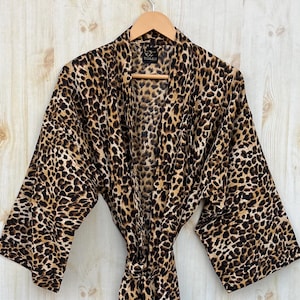 May include: A leopard print robe hanging on a wooden hanger. The robe is brown and tan with a repeating spotted pattern. It has wide sleeves and a matching belt. The robe is displayed against a light wood-paneled wall.