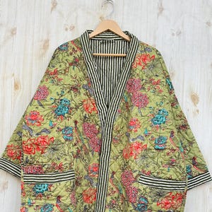 May include: A quilted, olive green robe with a floral pattern in red, blue, and pink. The robe has black and white striped trim and pockets. It is hanging on a wooden hanger.