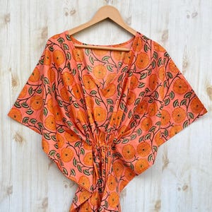 Puede incluir: Un caftán color melocotón con un estampado floral de flores naranjas y hojas verdes. El caftán tiene cuello en V y mangas anchas, y está colgado de una percha de madera. La tela es ligera y parece ser de algodón.