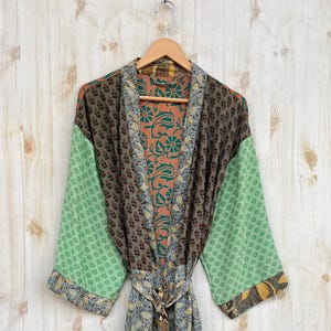 May include: A patchwork robe with a floral and geometric pattern in shades of green, brown, and blue hangs on a wooden hanger. The robe has long sleeves and a matching belt. The background is a light wood grain.