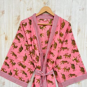 May include: A pink robe with a repeating pattern of brown tigers. The robe has a striped trim and a matching belt. The robe is hanging on a wooden hanger against a light wood background.