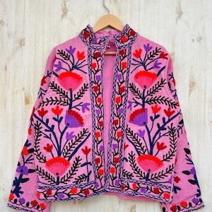 May include: A pink embroidered jacket with floral designs in red, blue, and purple. The jacket has a mandarin collar and long sleeves. The jacket is hanging on a hanger against a light wood background.