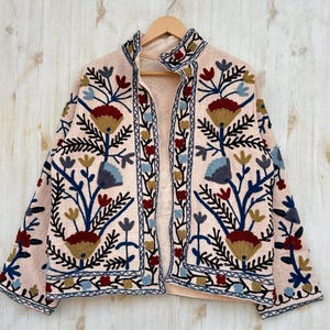 Pode incluir: Casaco bordado rosa claro com um design floral em tons de azul, vermelho e dourado. O casaco tem uma gola alta e est&aacute; pendurado em um cabide de madeira contra um fundo de madeira clara.