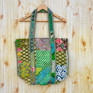 May include: A patchwork tote bag with green, blue, and white fabric. The bag has a long strap and is hanging on a wooden hanger.
