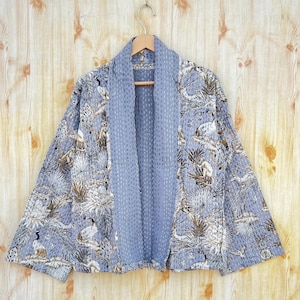 Puede incluir: Chaqueta kimono abierta azul claro con estampado botánico y de pájaros. La chaqueta acolchada tiene un forro azul claro liso. Cuelga de una percha de madera sobre un fondo de madera clara.