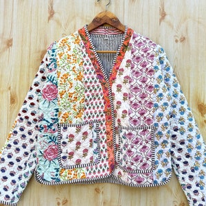 May include: A white quilted jacket with a patchwork design featuring floral and geometric patterns in shades of pink, orange, blue, and green. The jacket has a V-neck, long sleeves, and a single pocket on the front.