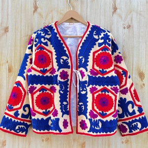 May include: A cream-coloured jacket with detailed embroidery in red, blue, and purple. The jacket has a floral design and a tie closure. It is displayed on a wooden hanger against a light wood backdrop.
