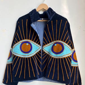 May include: A navy blue velvet jacket with embroidered eye designs. The jacket features light blue and brown eye details with gold sunburst rays. The interior lining has a blue and white striped pattern. The jacket is hanging on a wooden hanger.