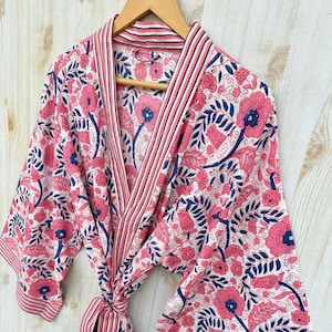 K&ouml;nnte beinhalten: Ein Bademantel mit Blumenmuster in Rosa, Wei&szlig; und Marineblau h&auml;ngt an einem Holzb&uuml;gel. Der Bademantel zeigt ein sich wiederholendes Muster aus gro&szlig;en rosa Blumen und blauen Bl&auml;ttern auf wei&szlig;em Grund. Er hat eine gestreifte Bord&uuml;re und eine passende Krawatte.