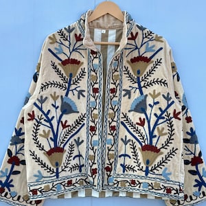 Pu&ograve; includere: Una giacca beige ricamata con un motivo floreale in tonalit&agrave; di blu, rosso e marrone. La giacca ha un colletto alto e una chiusura con cerniera sul davanti.