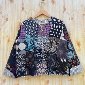 Peut inclure: Une veste matelassée noire avec un motif patchwork présentant divers motifs dans des tons de bleu, violet, blanc et noir. La veste a une fermeture boutonnée et une bordure rayée autour des bords.